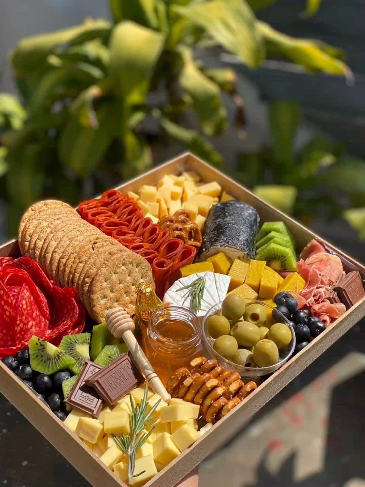 Caja de charcutería recién armada sostenida en alto: salami, galletas, cubos de queso, brie, aceitunas, arándanos, kiwi, chocolate y un frasco de miel con su cuchara, con vegetación desenfocada detrás.